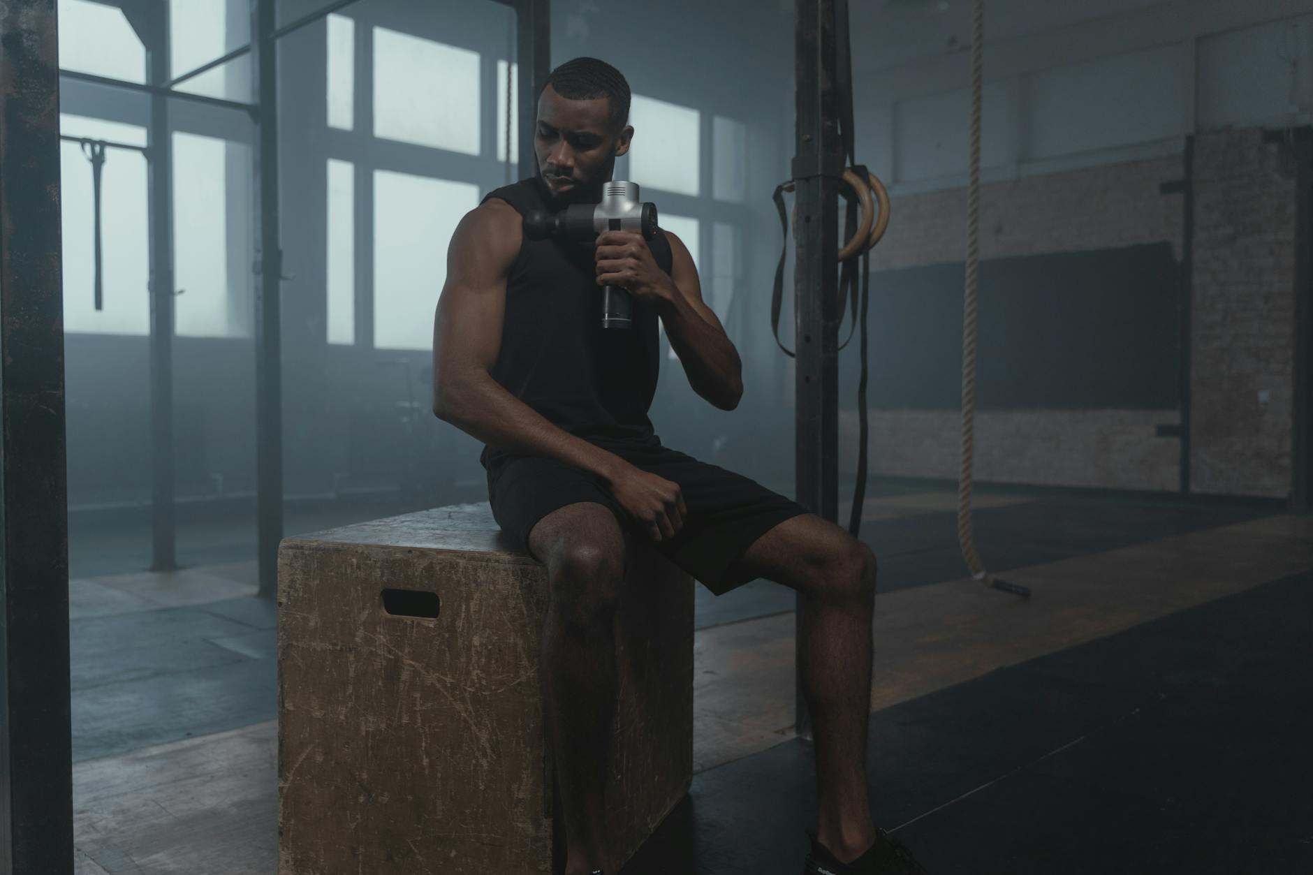 Fit man using a massage gun in a gym, wearing a tank top while seated on a wooden box.